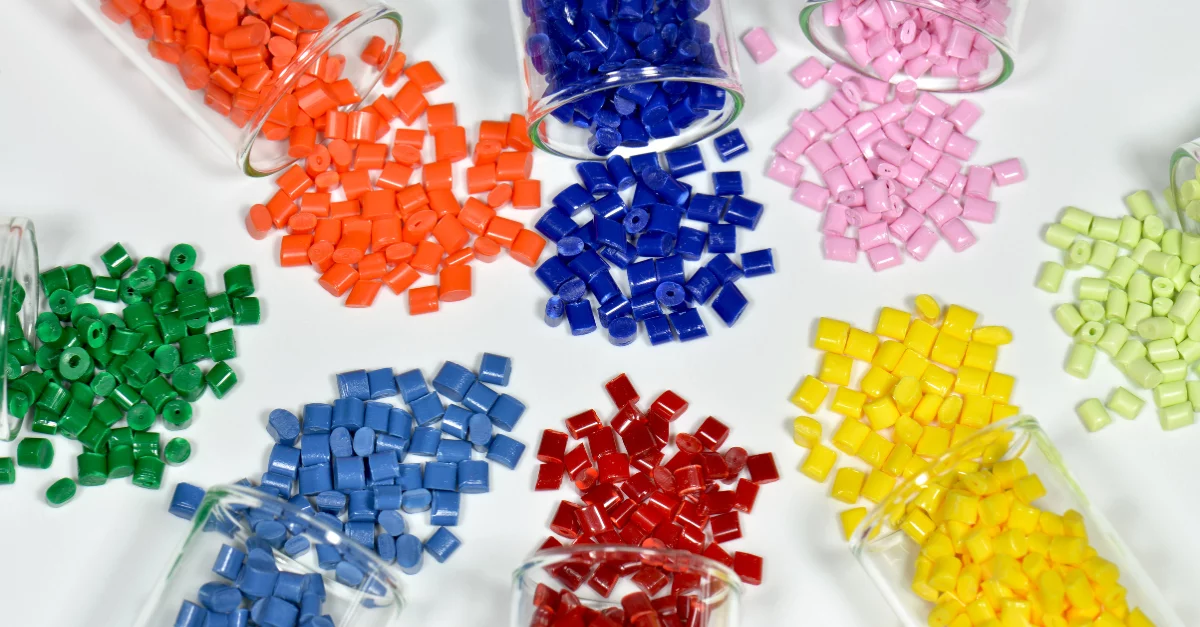 Vários tubos de ensaio de vidro contendo péletes de polímeros coloridos e translúcidos, dispostos em um rack de madeira em um laboratório. Uma variedade de cores vibrantes como vermelho, laranja, amarelo, rosa e azul é visível.
